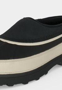 Chaussure en caoutchouc noire et blanche à enfiler, avec semelle épaisse crantée et bout arrondi, conçue pour les conditions extérieures ou humides.