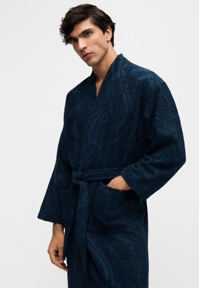 Jeune homme portant une robe de chambre texturée bleu foncé avec une ceinture, une main dans la poche, sur un fond clair uni.