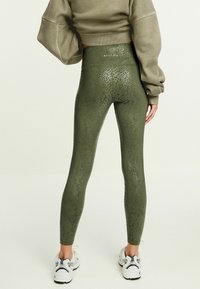 Olivgröna leggings med ett strukturerat mönster som liknar ormskinn, hög midja och en figurnära design, matchat med vita träningsskor.