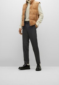 Chaleco acolchado marrón sobre un suéter color crema, combinado con pantalones gris oscuro y zapatos negros. Textura suave, botones a presión y detalle de bolsillo con cremallera.