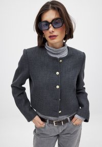 Tummanharmaa lyhythihainen bleiseri, jossa on pyöreä kaulus ja kultaiset napit. Päälle on puettu vaaleanharmaa poolopaita, yhdistettynä harmaisiin housuihin ja ruskeaan vyöhön.