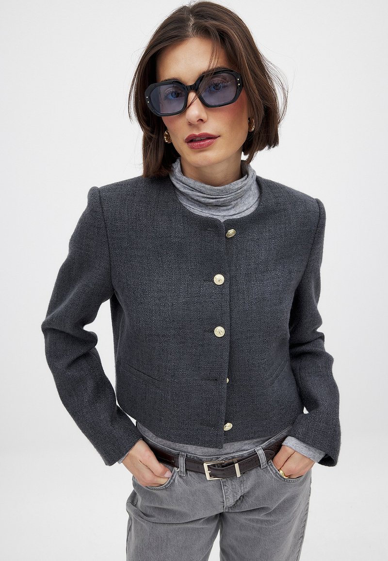 Tummanharmaa lyhythihainen bleiseri, jossa on pyöreä kaulus ja kultaiset napit. Päälle on puettu vaaleanharmaa poolopaita, yhdistettynä harmaisiin housuihin ja ruskeaan vyöhön.