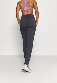 Mujer vistiendo un sujetador deportivo mauve, pantalones jogger gris oscuro y zapatillas blancas con acentos rosas, de pie contra un fondo blanco liso.