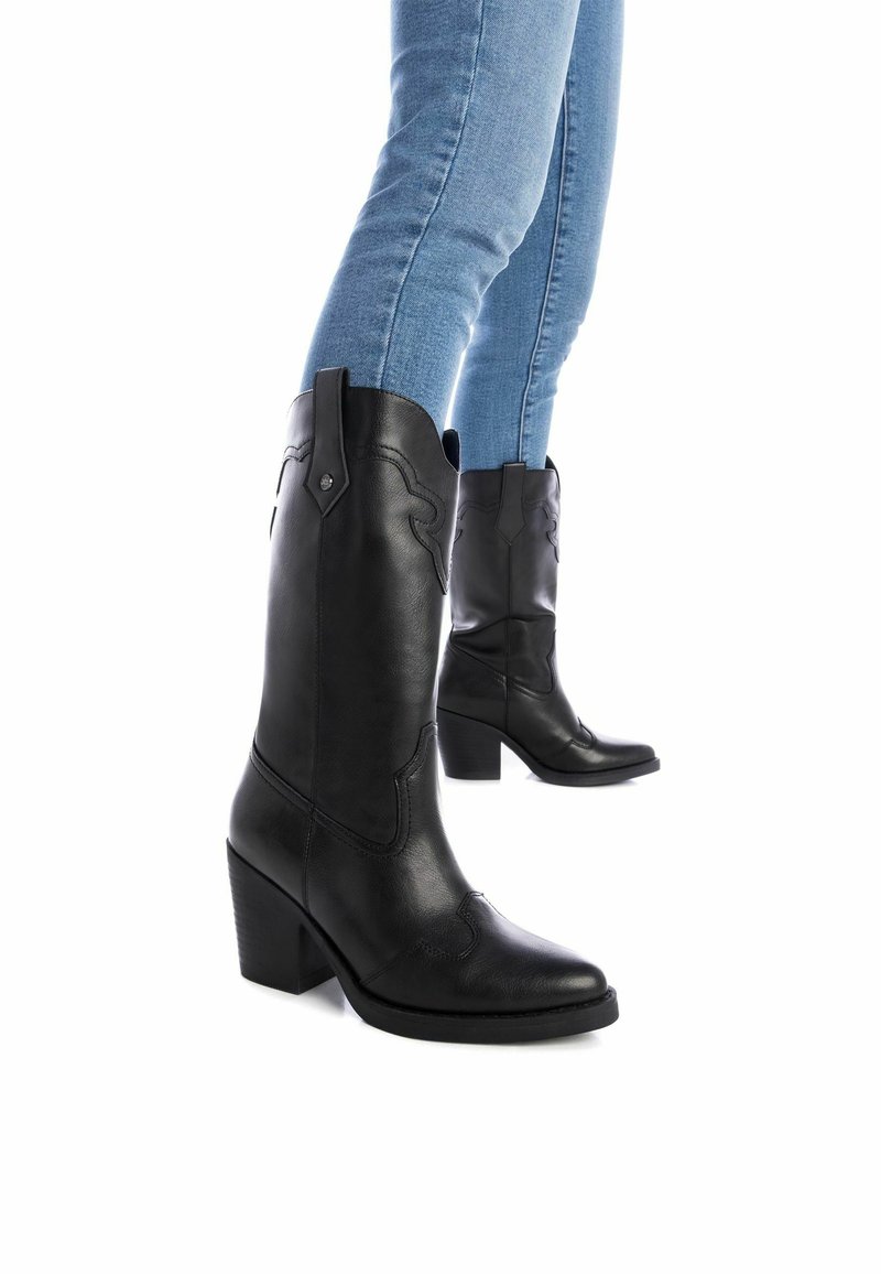 Botas de vaquero de cuero negro con punta afilada, diseño de costura intrincado y tacón grueso, combinadas con jeans ajustados de color azul claro.