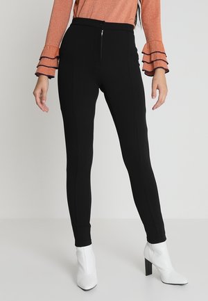 Woman wearing black slim pants, orange long-sleeve top with ruffled cuffs, and white high-heeled ankle boots standing on a white floor.