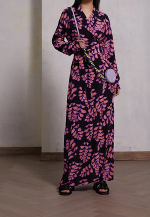 Kobieta w czarnej długiej sukni maxi z pomarańczowo-fioletowymi wzorami liści, niosąca małą, okrągłą lawendową torebkę na ramię z plecionym paskiem.