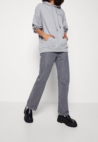 Sudadera con capucha gris, con bolsillo frontal, cordones y detalle de logo, combinada con jeans gris de pierna recta y zapatos negros gruesos.