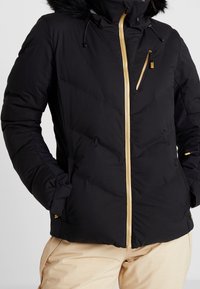 Doudoune noire avec fermeture éclair à l'avant, accents dorés et finition texturée. Comprend une capuche amovible doublée de fourrure et une poche poitrine zippée.
