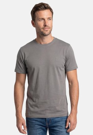 Mies, jolla on lyhyet ruskeat hiukset, pukeutuneena harmaaseen lyhythihaiseen t-paitaan ja sinisiin farkkuihin, katsoo hieman sivulle ilmeettömänä.