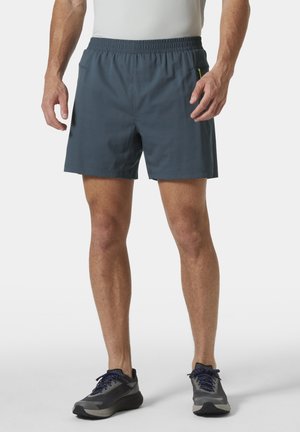 Mężczyzna w prostych ciemnoszarych sportowych szortach z elastycznym pasem i kieszenią na zamek, w połączeniu z szaro-niebieskimi butami do biegania.