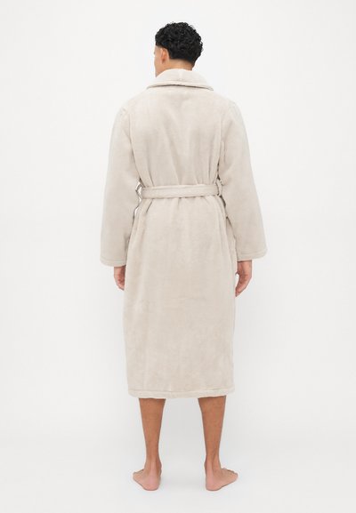 Roupa de banho bege e felpuda com colarinho tipo xale, mangas longas e cinto amarrado na cintura, apresentando uma superfície texturizada e design até ao joelho.
