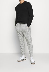 Zwart textuurtrui, grijs geruite tapered broek met koord, en witte sneakers met zwarte strepen. Model staat tegen een effen achtergrond.