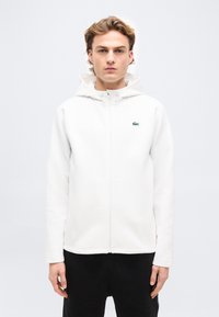 Νεαρός άνδρας με ανοιχτό καστανά μαλλιά, που φοράει λευκό φούτερ με φερμουάρ και κουκούλα, καθώς και μαύρο παντελόνι, στέκεται μπροστά σε απλό ανοιχτόχρωμο φόντο.