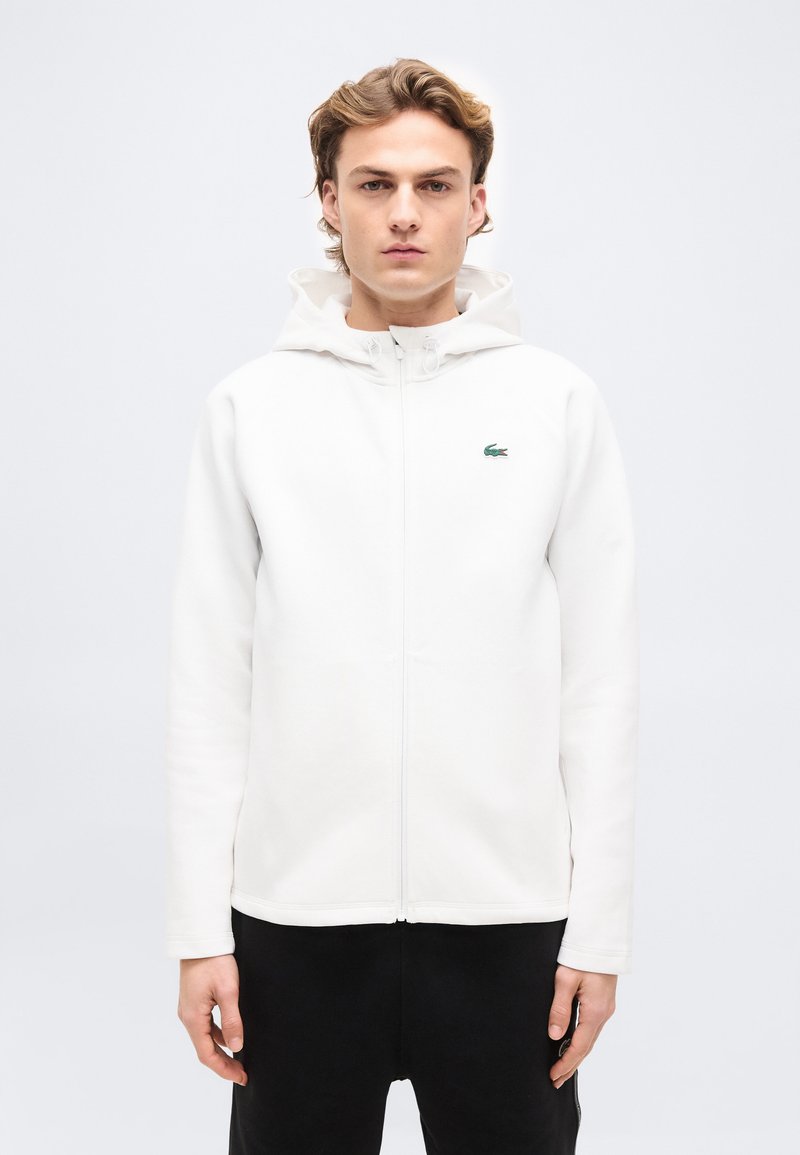 Νεαρός άνδρας με ανοιχτό καστανά μαλλιά, που φοράει λευκό φούτερ με φερμουάρ και κουκούλα, καθώς και μαύρο παντελόνι, στέκεται μπροστά σε απλό ανοιχτόχρωμο φόντο.