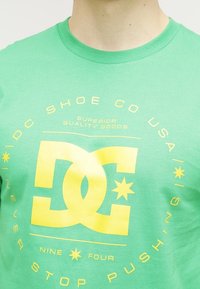 Zelené bavlněné tričko má velký žlutý grafický motiv s textem, včetně "DC SHOE CO USA" a stylizovaného loga ve středu.