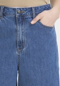 Jean en denim bleu avec une coupe droite, doté d'une poche avant, d'une fermeture par bouton et de coutures contrastantes le long des coutures.