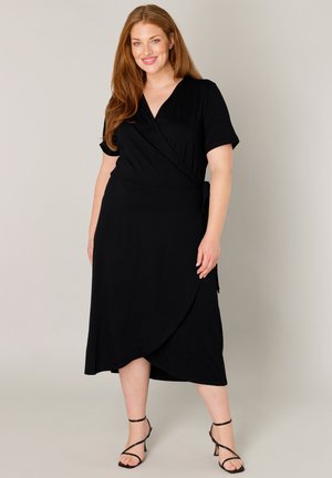 Plus-size vrouw met lang rood haar, gekleed in een zwarte wikkeljurk en zwarte sandalen met bandjes, glimlachend tegen een neutrale achtergrond.