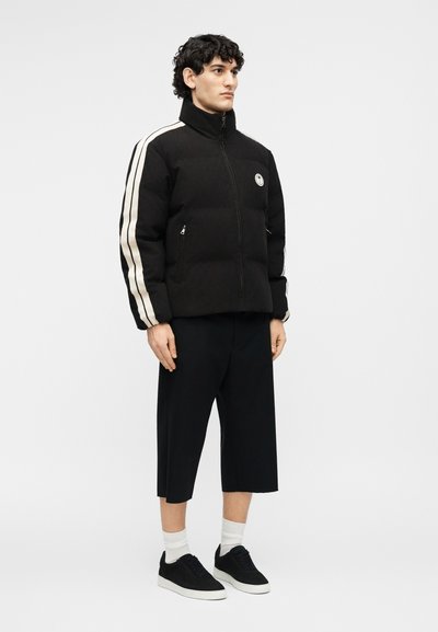 Άνδρας που φοράει μαύρο μπουφάν puffer με λευκές ρίγες στα μανίκια, μαύρο cropped παντελόνι, λευκές κάλτσες και μαύρα αθλητικά παπούτσια, στέκεται μπροστά σε απλό φόντο.