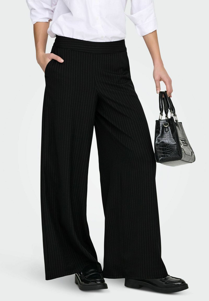 Černé kalhoty s širokými nohavicemi a jemným proužkem, s hladkým pasem; doplněné malou černou texturovanou kabelkou a lesklými černými botami.