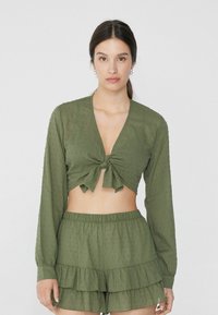 Blusa corta verde oliva con maniche lunghe e dettaglio legato davanti, abbinata a pantaloni corti a balze. La texture presenta strisce verticali e piccoli punti.