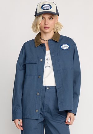 Marineblauwe werkjas met een bruine corduroy kraag, voorzakken en drukknoopsluiting; gecombineerd met een bijpassende blauwe outfit en een pet met een logo.