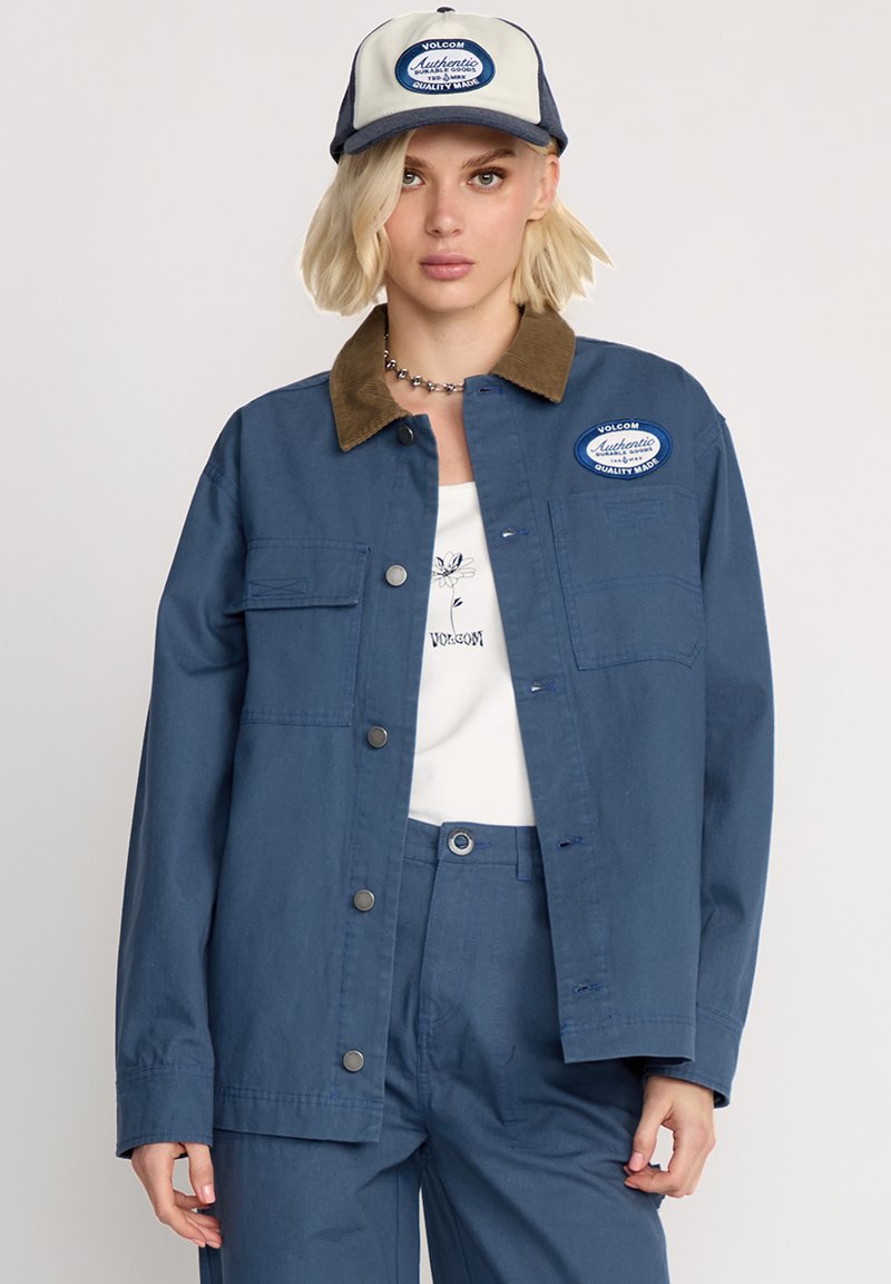 Marineblaue Arbeitsjacke mit einem braunen Cordkragen, Fronttaschen und Druckknöpfen; kombiniert mit einem passenden blauen Outfit und einer Kappe mit einem Logo.