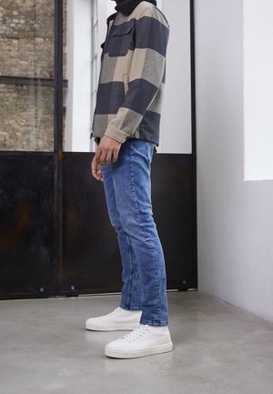 Persoon die een blauwe spijkerbroek, witte hoge sneakers en een beige en donkergrijs geruit jack draagt, staat binnen op een betonnen vloer.