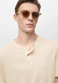 Světle béžová henley košile s třemi knoflíky, hladká textura a zaoblený výstřih. Model má na sobě sluneční brýle s amber skly a průhledným rámem.