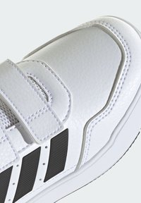 Biela teniska s textúrovaným syntetickým zvrškom, suchým zipsom a čiernymi pruhmi. Stehovanie zvýrazňuje dizajn.