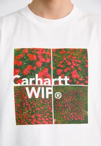 Biela tričko s štvorcovým grafickým motívom so štyrmi obrázkami červených kvetov na zelenej tráve, prekryté nápisom "Carhartt WIP®" tučným písmom.
