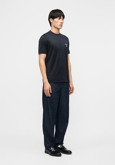 Νέος άνδρας που στέκεται γυρίζοντας προς τα δεξιά, φοράει μαύρο μπλουζάκι με λογότυπο στο στήθος, σκούρο τζιν με φαρδιά πόδια και μαύρα δερμάτινα παπούτσια, μπροστά σε απλό φόντο.