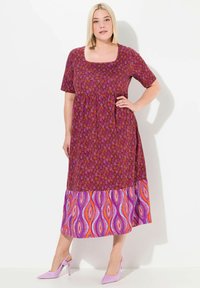 Lila und rosa gemustertes Maxikleid mit quadratischem Ausschnitt, kurzen Ärmeln und einem lebhaften gemusterten Saum. Kombiniert mit helllila Absätzen.