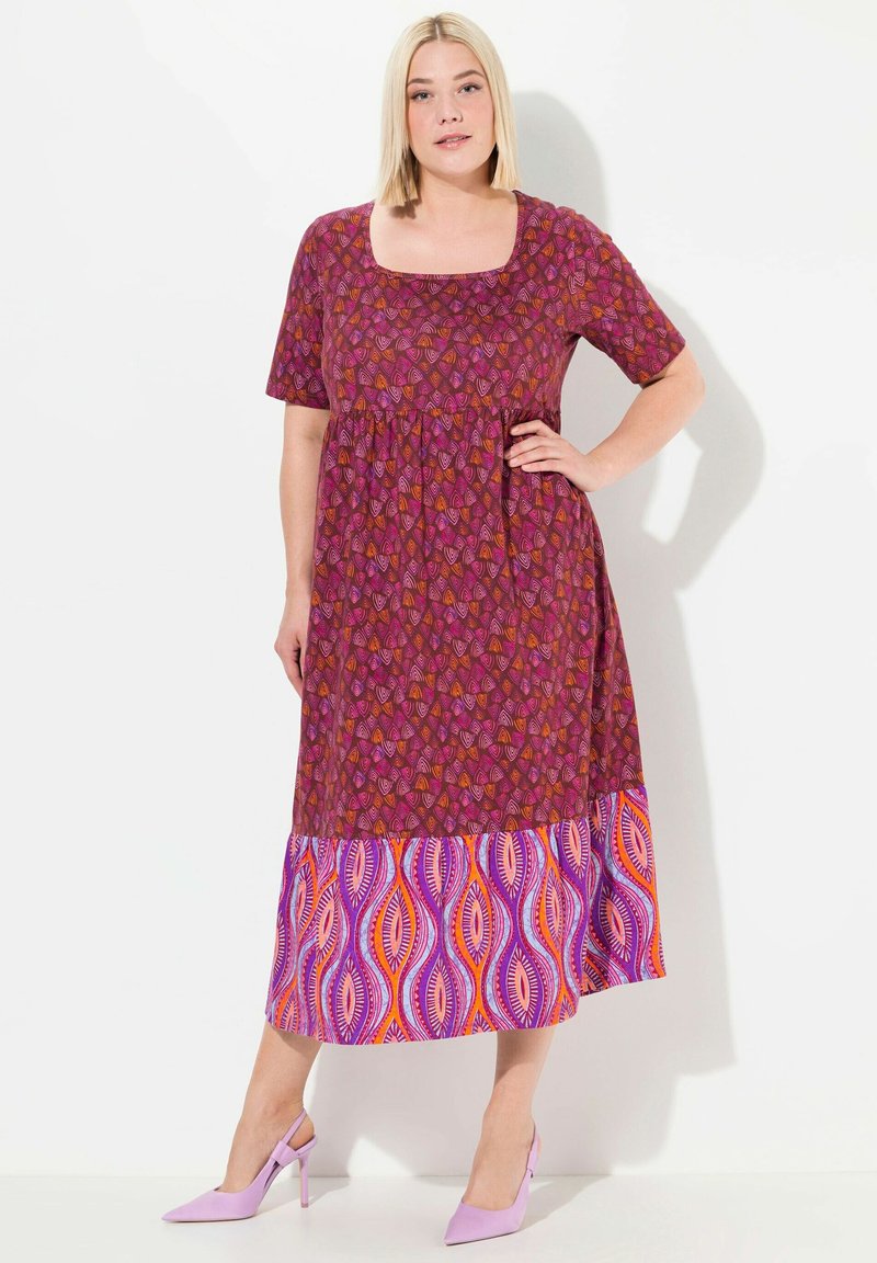 Lila und rosa gemustertes Maxikleid mit quadratischem Ausschnitt, kurzen Ärmeln und einem lebhaften gemusterten Saum. Kombiniert mit helllila Absätzen.