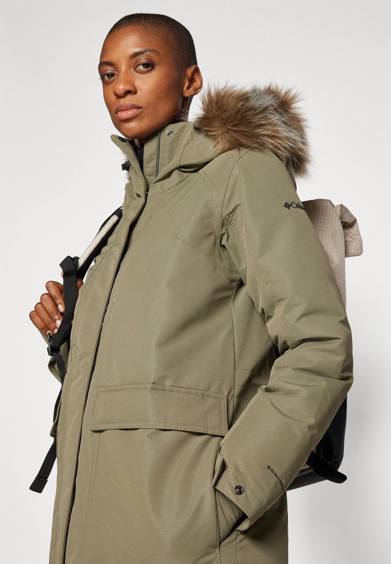Olivgrüner isolierter Parka mit einer mit Fell gefütterten Kapuze, ausgestattet mit Reißverschlusstaschen und verstellbaren Bündchen, getragen über einem dunklen Oberteil und einem Rucksack.