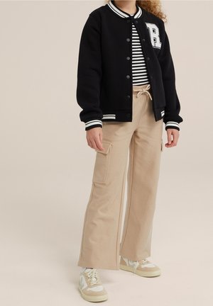Veste universitaire noire avec des poignets à rayures blanches, associée à une chemise à rayures noir et blanc et à un pantalon cargo beige. Baskets avec des accents crème.
