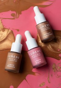 Drie glazen pipetflessen: bruin voor Liquid Bronzer, roze voor Liquid Blush, en brons voor Instant Illuminizer, gezet op een kleurrijke achtergrond.