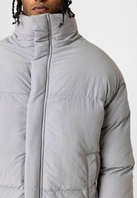 Lichtgrijze pufferjack met een hoge kraag, met een gladde textuur, gestikte naden en een voorzak voor opbergruimte.