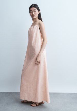 Mladá žena s dlhými tmavými vlasmi nosí voľné, bezrukávové maxi šaty s ružovo-bielymi pruhmi a hnedé ploché sandále, stojaca zboku.