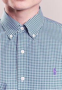 Grön- och vitrutigt knäppskjorta med klassisk krage och vita knappar; har en liten lila logotyp på bröstet.