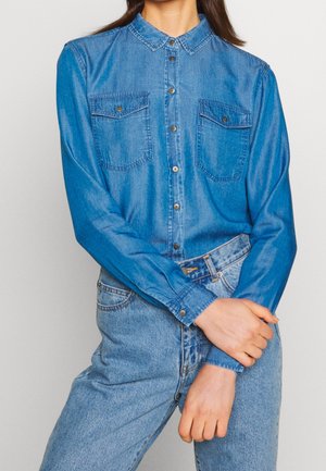 Personne portant une chemise en denim bleu à boutons avec deux poches poitrine et un jean taille haute bleu clair, les mains croisées devant.