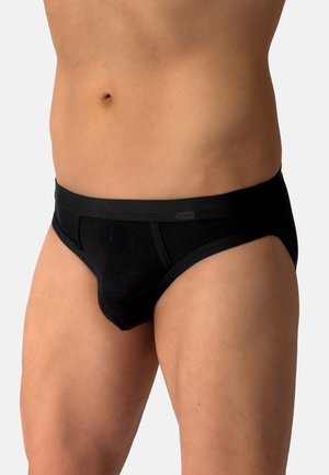 Männlicher Oberkörper mit schwarzer Slip-Unterwäsche vor einem schlichten hellen Hintergrund, zeigt Taille, Hüften und obere Oberschenkel.