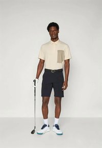 Camisa polo bege com um bolso de remendo verde, combinada com calções azul-marinho. Figura em pé segura um taco de golfe, vestindo sapatos brancos com detalhes azuis.