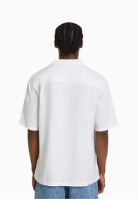 Person med flätat hår som bär en enkel vit kortärmad skjorta och blå denimshorts, visad bakifrån mot en vit bakgrund.