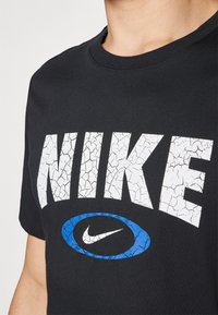 Crni pamučni majica s velikim, napuknutim bijelim logotipom "NIKE" i plavim ovalnim naglaskom unutar kojeg se nalazi Nike Swoosh simbol.