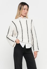 Witte gebreide trui met zwarte verticale accenten en een kabelgebreide structuur. Wijde mouwen en geribbelde zoom, gedragen over een witte blouse met kraag.