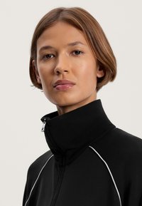 Fekete sportos dzseki magas nyakkal, cipzárral, és fehér díszítéssel a vállakon. Simára szőtt anyag.