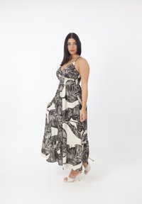 Mujer con cabello largo y oscuro, vestida con un vestido maxi sin mangas en blanco y negro con estampado de hojas, y sandalias blancas de tacón alto, posando de perfil.