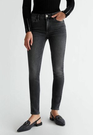 Kvinde iført sort ribbet langærmet top med naglede manchetter, tætsiddende falmede sorte skinny jeans og sorte spidse slingback flade sko med spænde.