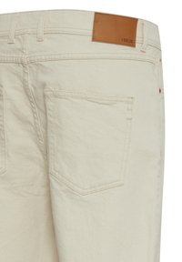 Pantalones de algodón beige con superficie texturizada, que cuentan con dos bolsillos traseros, un parche de cuero marrón en la cinturilla y detalles de costura mínimos.