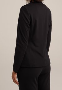 Person med lockigt hår som bär en figursydd svart kavaj och svarta byxor, sedd bakifrån mot en enfärgad bakgrund.